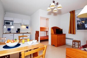 A compact room with a kitchenette, a dining table with fruit and a glass of juice, a bunk bed, a TV, and a window with curtains. - Studio au calme avec accès piscine (Font Romeu)