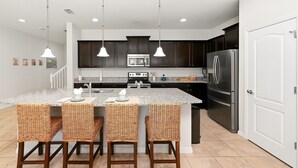 A kitchen with dark cabinets, a stainless steel refrigerator, a microwave, and a granite countertop. There are three wicker chairs around the island. - Group Hub Near Disney | 4BR with Pool and BBQ | Windsor of Westside Resort (Four Corners)