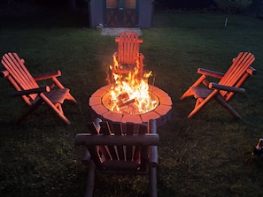 A circular fire pit with logs, surrounded by four Adirondack chairs.