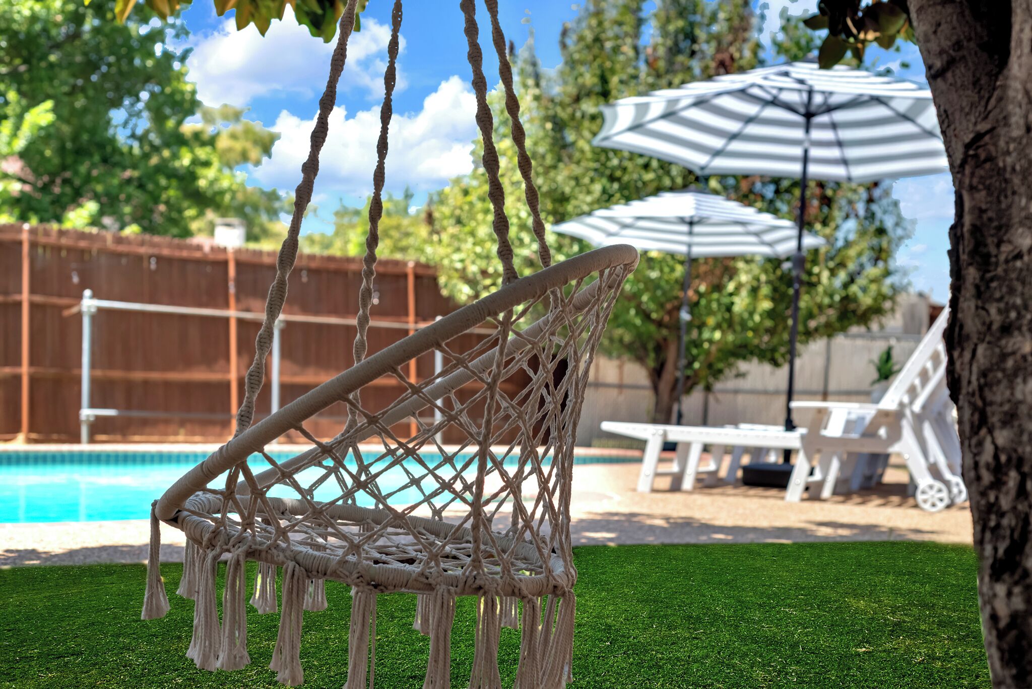 A hammock chair with a netted design, a pool, and a white umbrella in a backyard setting.