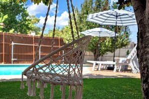 Una silla hamaca con diseño de red, una piscina y una sombrilla blanca en un patio.