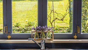 Una cocina con un lavamanos de acero inoxidable, una ventana con vista a un jardín y un estante de madera con jarrones.