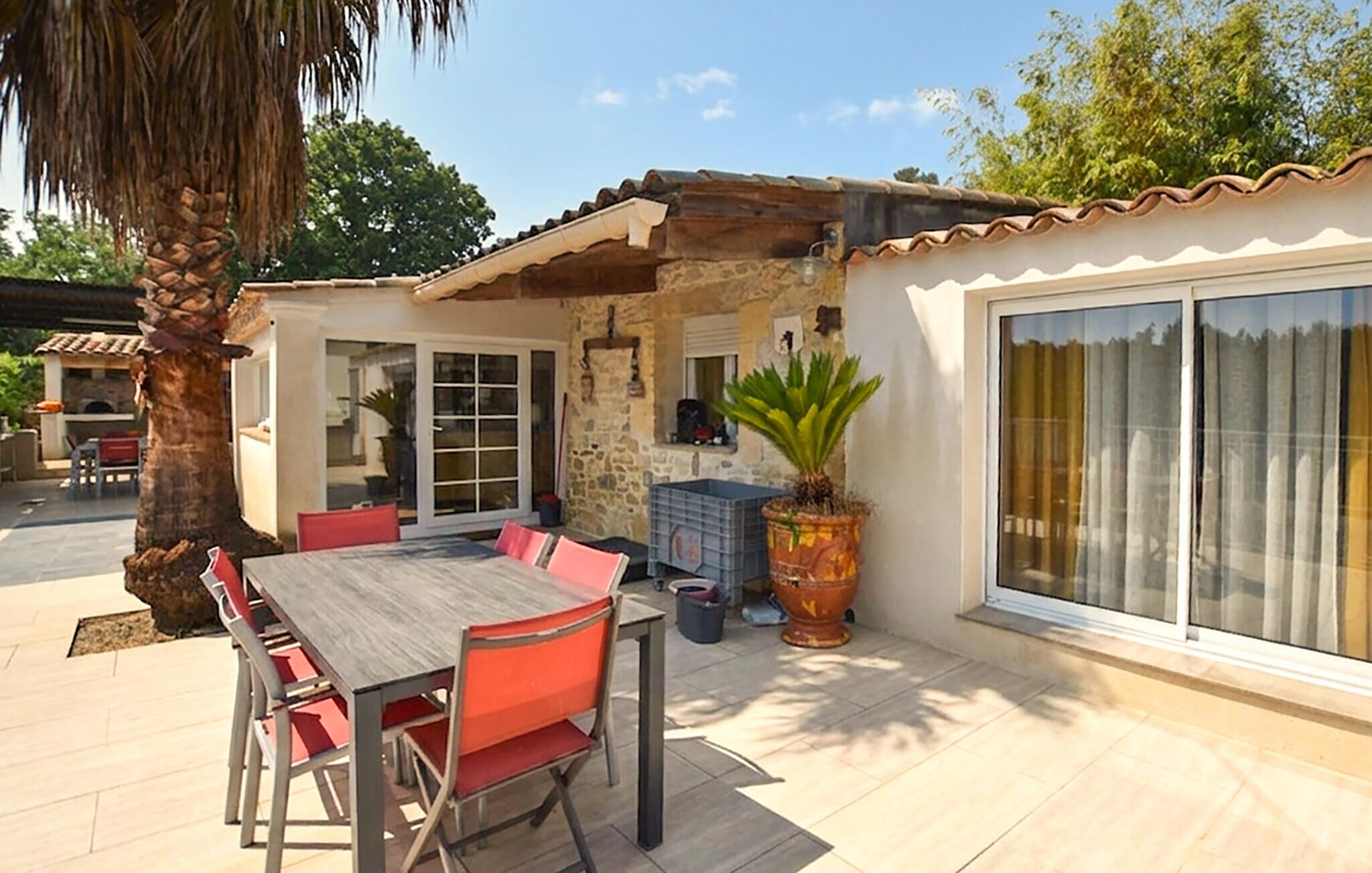 Un patio esterno con tavolo di legno e sedie rosse, un muro in pietra e un'ampia finestra con tende.