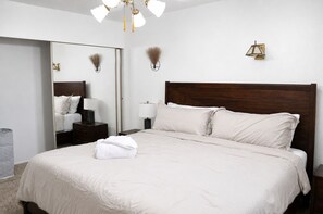 Uma cama bem arrumada com edredom branco e almofadas, uma mesa de cabeceira com abajur, um espelho fixado na parede e um arandela.