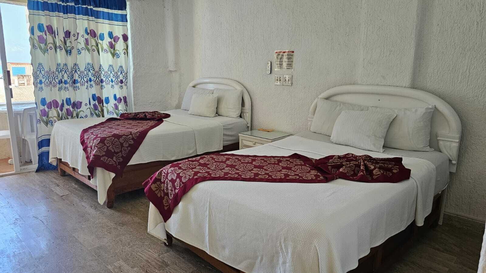 Due letti singoli con lenzuola bianche e coperte a fiori rossi in una stanza con una parete bianca e una finestra con tenda floreale.