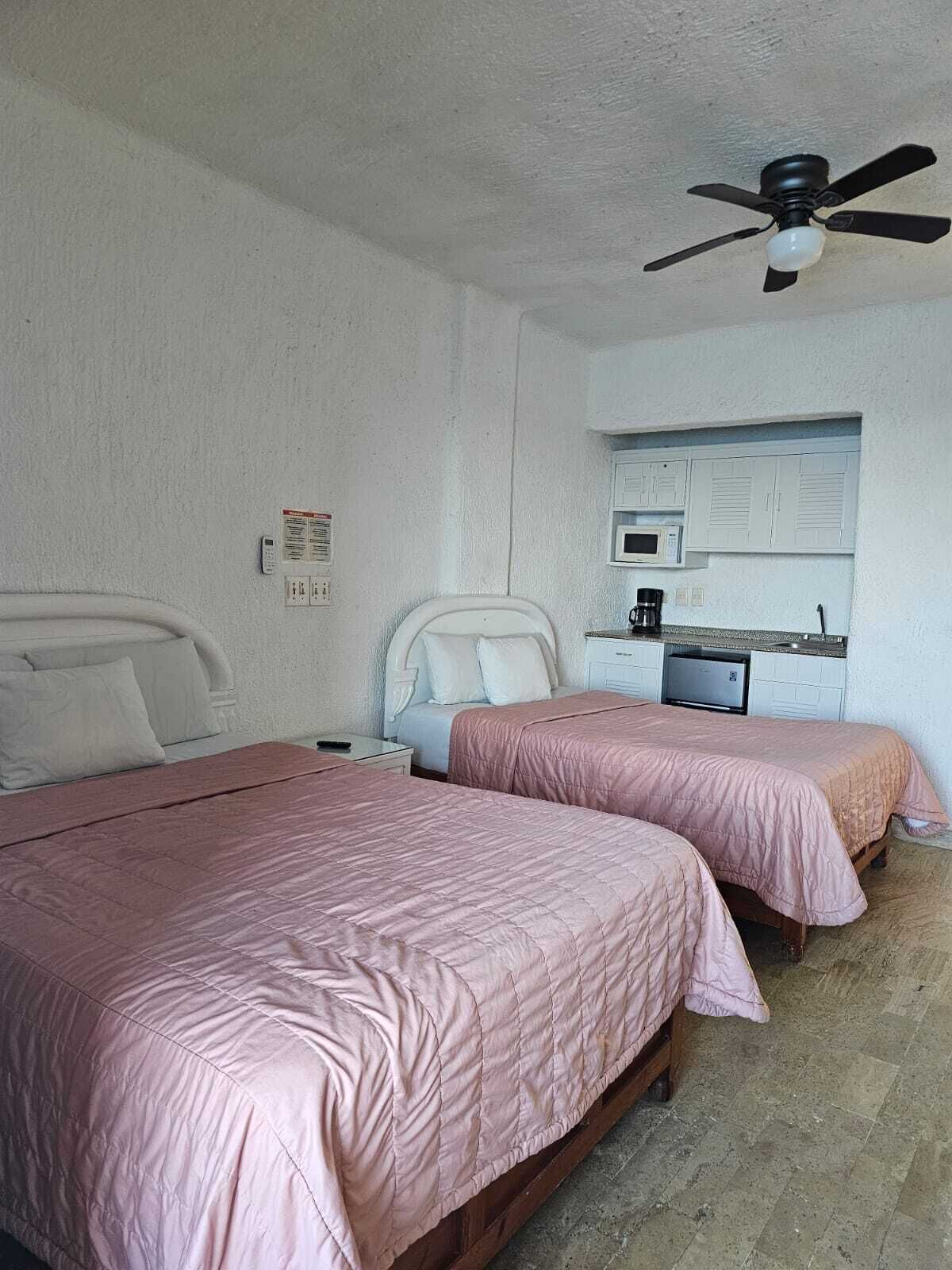 Une chambre avec deux lits, un ventilateur de plafond, un coin cuisine et un mur blanc.