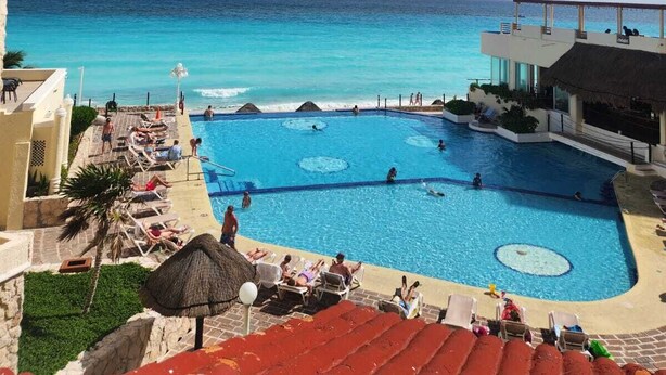 Piscina de resort com águas azuis cristalinas, rodeada por espreguiçadeiras e guarda-sóis, com vista para uma praia e oceano.