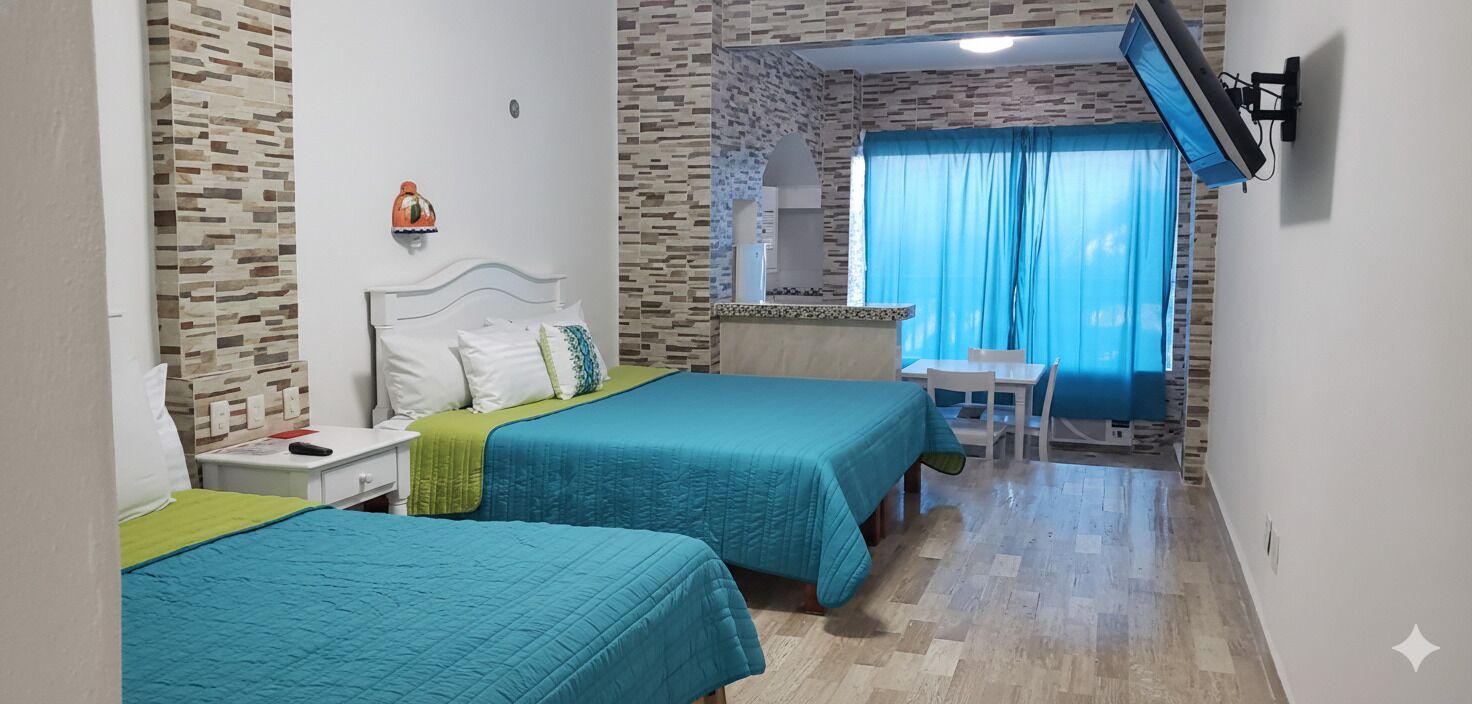 A hotel room with two beds, a small kitchenette, a TV mounted on the wall, and a window with curtains.