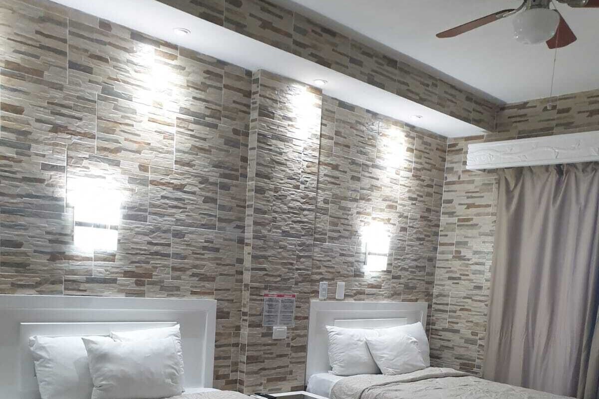 Quarto de hotel com duas camas, um ventilador de teto e parede com padrão de pedra.