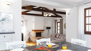 Ein Essbereich mit Holztisch, weißen Stühlen, Kamin und einer Couch im Hintergrund.