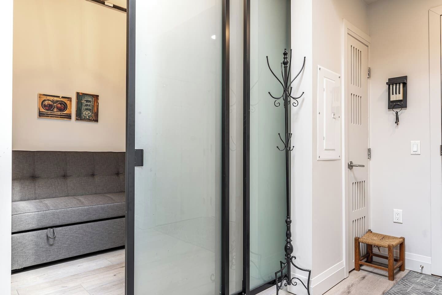 A modern interior with a grey sofa, a glass partition, a coat rack, and a small wooden stool.
