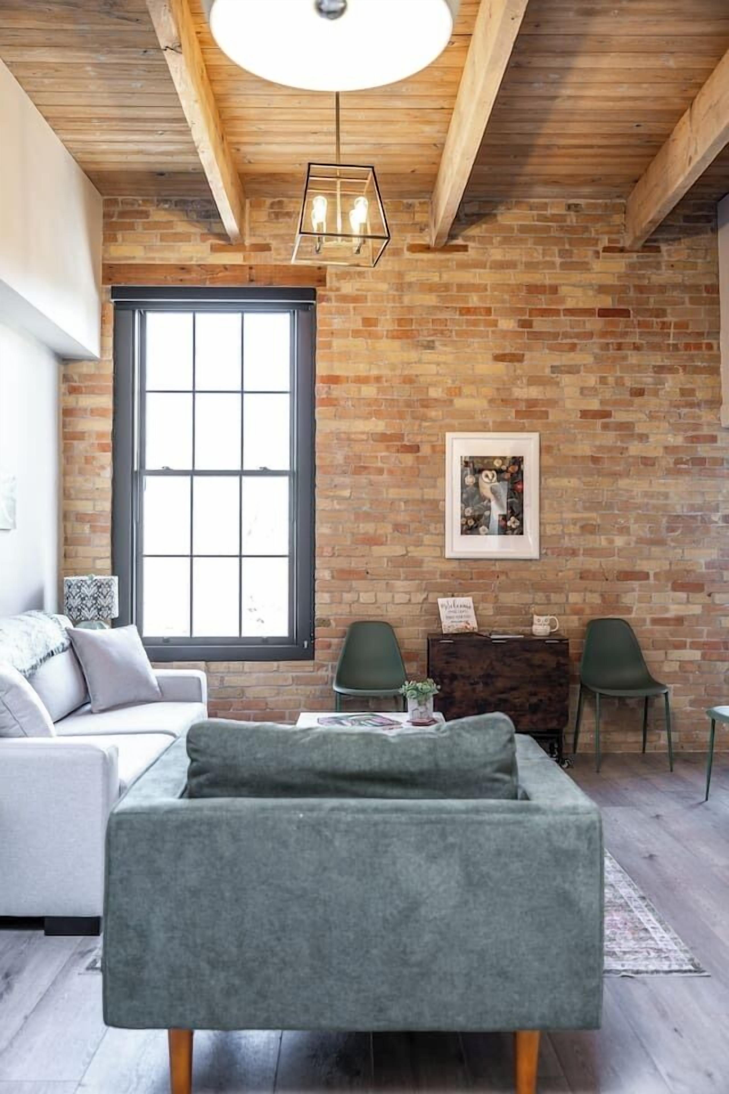 Une pièce aux murs en briques apparentes, avec une grande fenêtre, un canapé, deux fauteuils, une table et un luminaire suspendu.