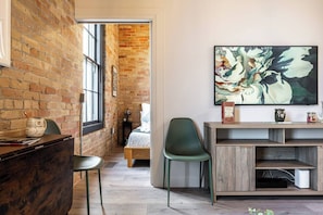 A room with a brick wall, a large abstract painting, a desk with a chair, and a bed with a green chair.