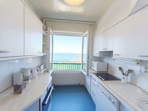 A compact kitchen with white cabinets, a microwave, a kettle, and a view of the sea through an open door.