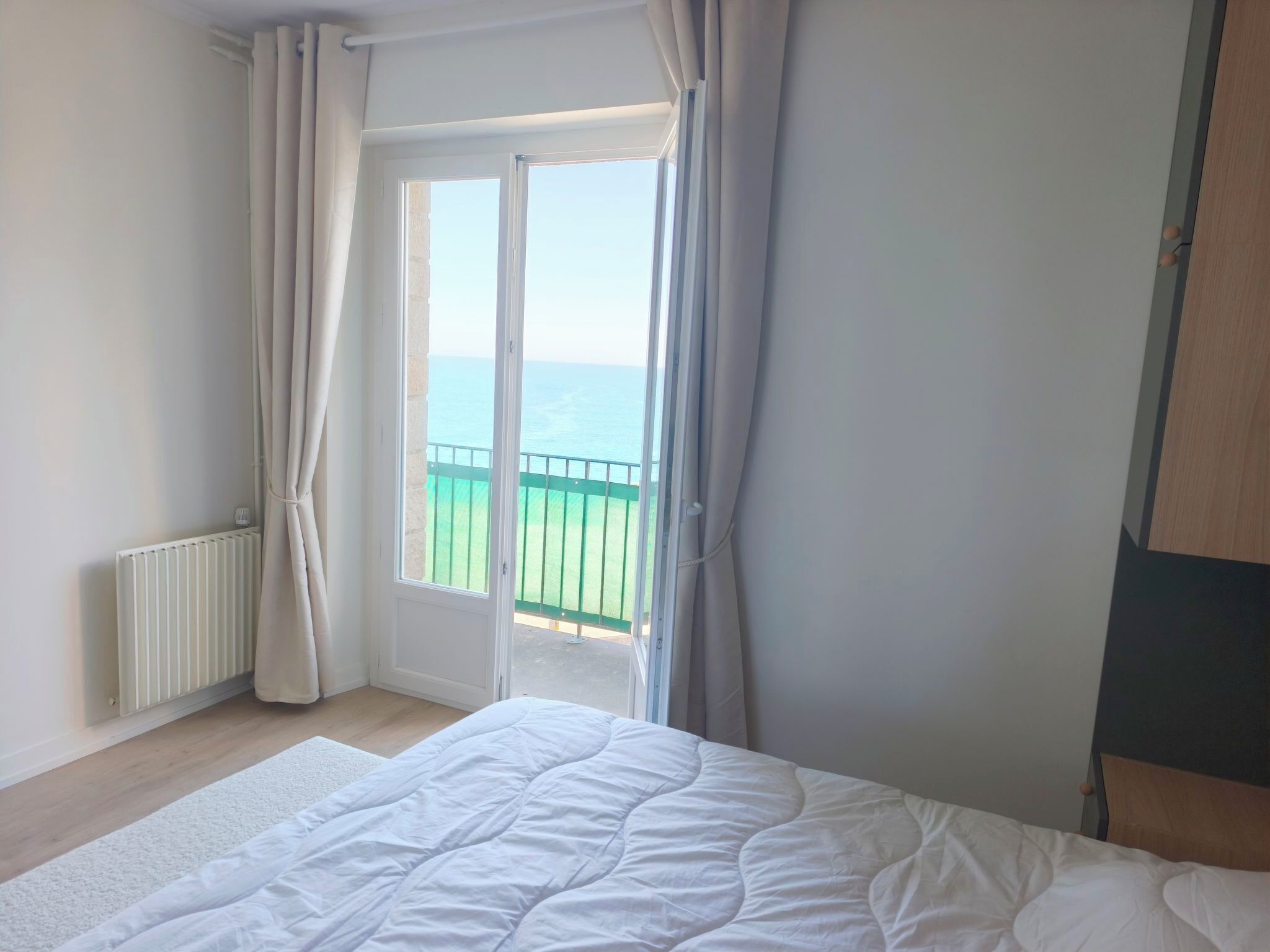 Ein Schlafzimmer mit Bett, Heizkörper, einem Fenster mit Vorhängen und Blick aufs Meer.