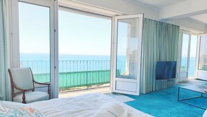 A hotel room with a bed, a chair, a TV, and a balcony with a view of the sea.