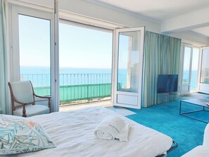 A hotel room with a bed, a chair, a TV, and a balcony with a view of the sea.