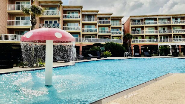 A swimming pool with a mushroom-shaped water feature, surrounded by multi-story hotel buildings with balconies. - The Jellyfish Resort-style (Galveston)