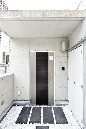Ein Gebäude mit Betonfassade, ein Eingangsbereich mit einer Tür, auf der 'Benvenuto 10' steht, ein Metallrahmen und ein Kiesweg, der zur Tür führt.