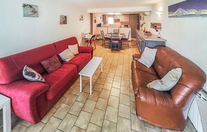 A living room with a red sofa, a brown leather sofa, and a white table. There is a kitchen in the background with a dining table and chairs.