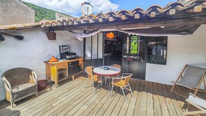 Um deck de madeira com mesa e cadeiras, uma área de cozinha com fogão e eletrodoméstico com a marca Castrol, e vista para uma encosta verde.