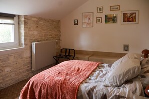 A bedroom with a stone wall, a radiator, a bed with a patterned blanket, a chair, and several framed pictures on the wall. - Au Logis Noisette - SAINT LOUP GEANGES (SAINT LOUP GEANGES)