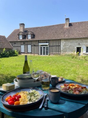 Ein Tisch im Freien mit Speisen und Getränken, im Hintergrund ein Haus mit Steinmauern und einem Ziegeldach.