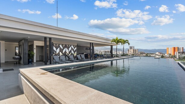 A rooftop infinity pool with lounge chairs, a modern building with geometric wall design, and a clear view of distant buildings and mountains. - Villa Borghese Roma · Condo in Trendy Versailles (Puerto Vallarta)