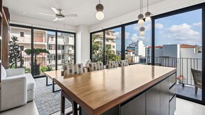 A modern kitchen with a large island, a dining area with chairs, a ceiling fan, and large windows offering a city view. - Villa Borghese Roma · Condo in Trendy Versailles (Puerto Vallarta)
