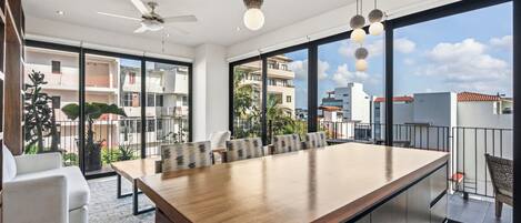 Una cocina moderna con una isla grande, un comedor con sillas, un ventilador de techo y ventanas amplias con vistas a la ciudad.