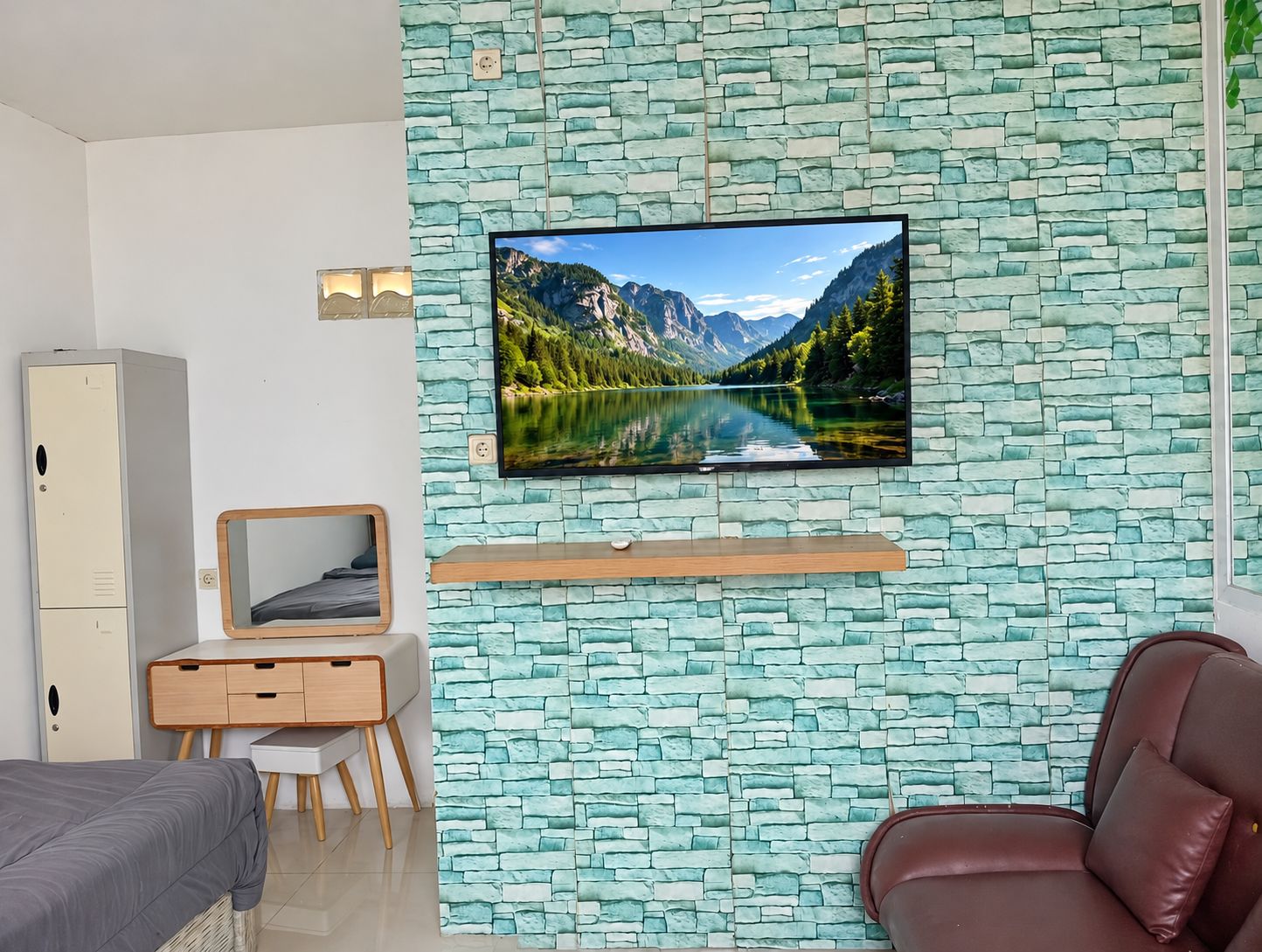 Un dormitorio con pared de diseño de piedra, un televisor montado, una cama, un escritorio con silla y un sofá de cuero marrón.