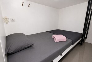 A single bed with a grey mattress and a pink towel, a power outlet, and two wall-mounted hooks.