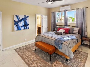A bedroom with a bed, a bench, a rug, an air conditioning unit, and a window with curtains.