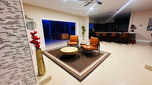 Ein modernes Wohnzimmer mit einem runden Couchtisch, orangefarbenen Sesseln, einer Vase mit roten Blumen, einem Deckenventilator und Blick in den Küchenbereich.