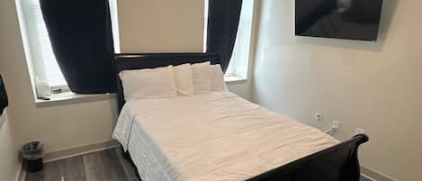 A bedroom with a bed, two windows with curtains, a flat-screen TV on the wall, and a waste bin.