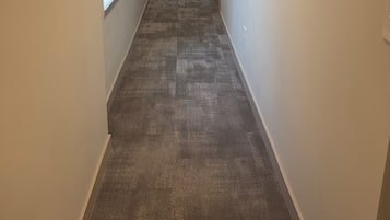 Un estrecho pasillo con pisos alfombrados, paredes blancas y una puerta al final que da a una habitación con cama y cómoda.