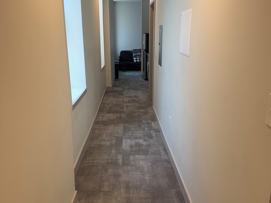 Un estrecho pasillo con pisos alfombrados, paredes blancas y una puerta al final que da a una habitación con cama y cómoda.
