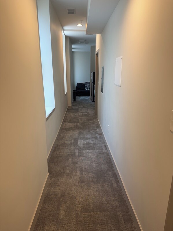 A narrow hallway with carpeted floors, white walls, and a door at the end leading to a room with a bed and a dresser.