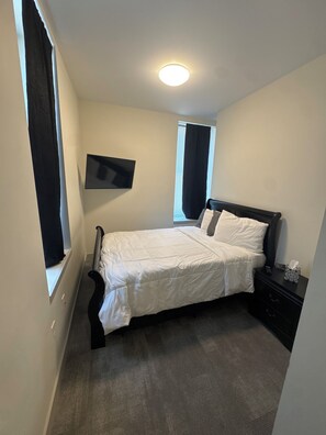 A bedroom with a bed, a nightstand, a TV mounted on the wall, and a window with curtains.