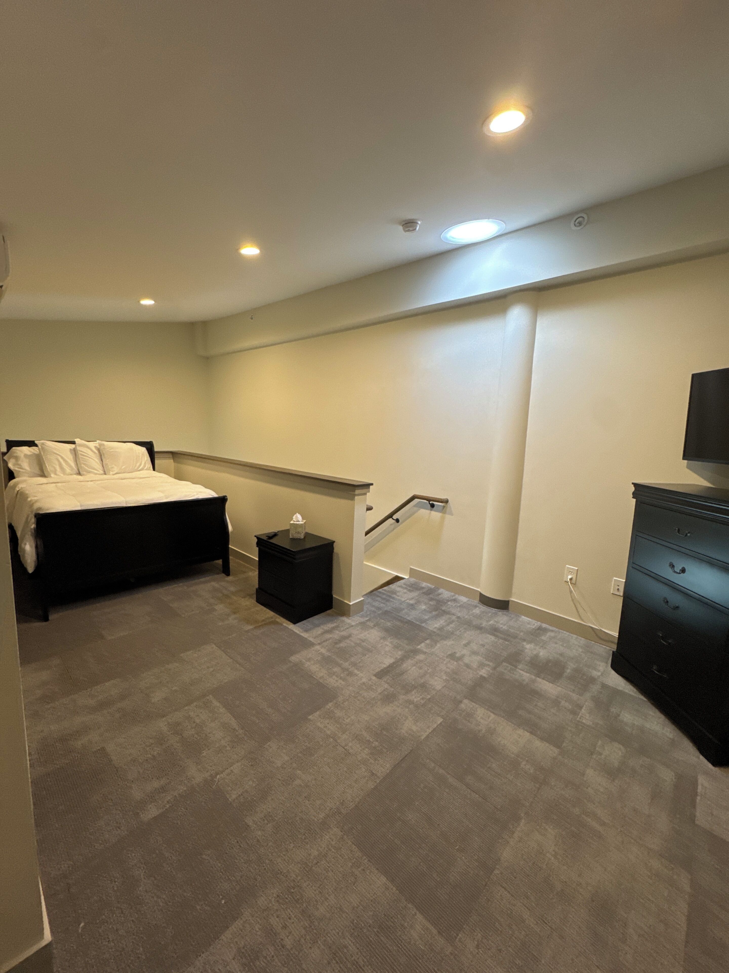 Un dormitorio con cama, mesita de noche, cómoda, escalera y televisor en la pared.