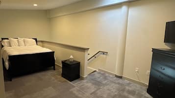 Un dormitorio con cama, mesita de noche, cómoda, escalera y televisor en la pared.