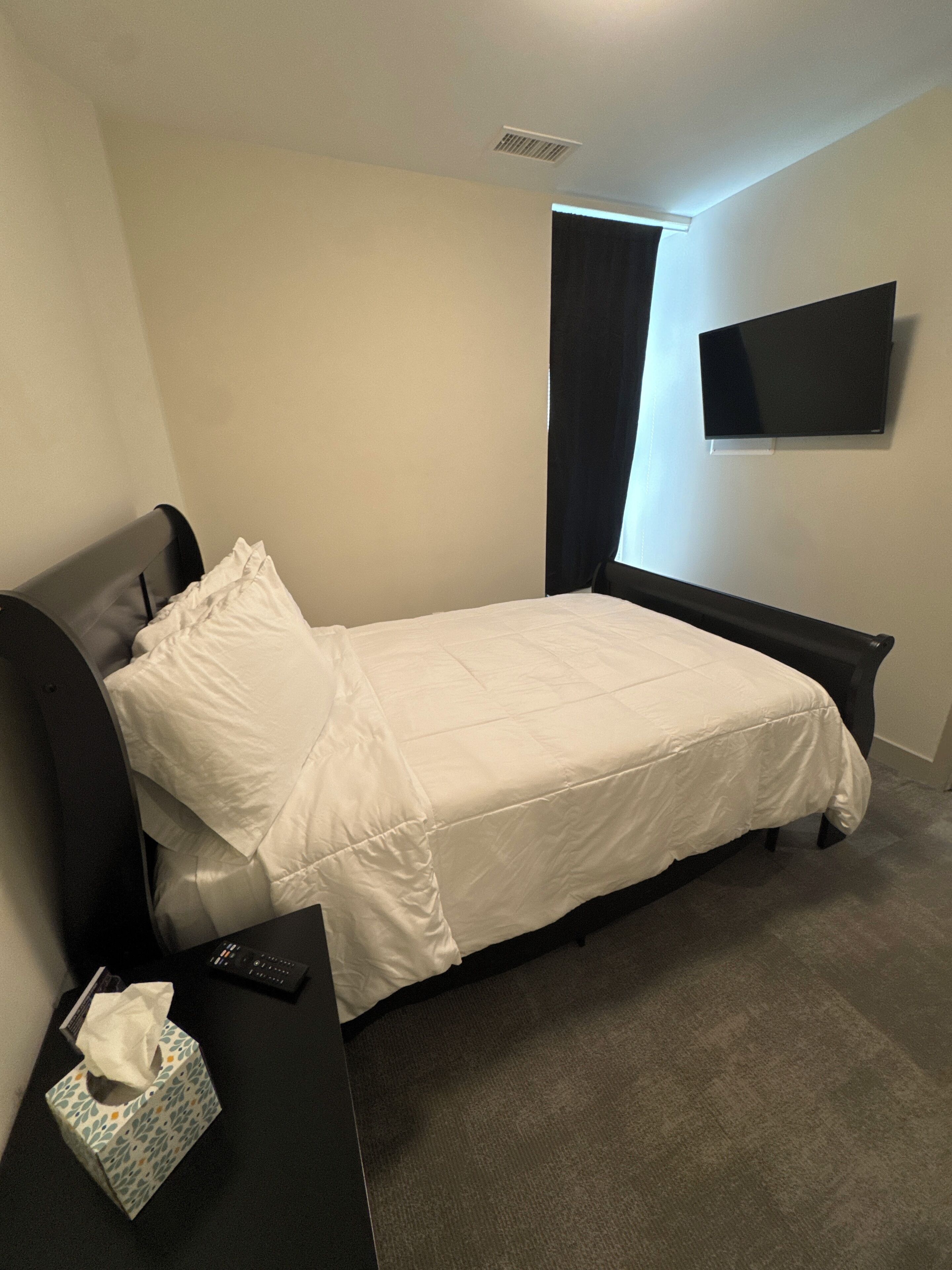 A single bed with a white comforter, a black headboard, a flat-screen TV on the wall, and a bedside table with a tissue box.