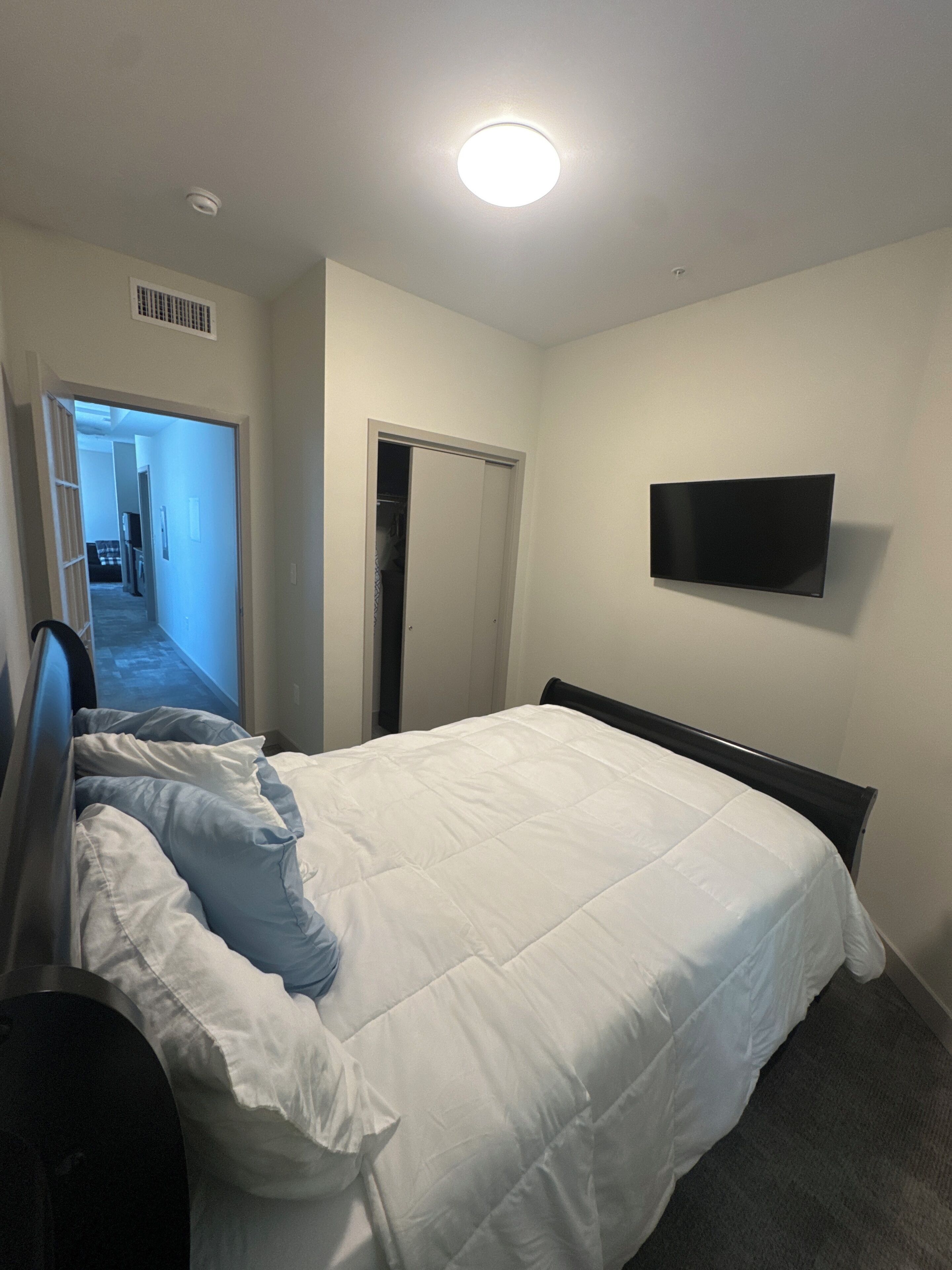 A bedroom with a bed, a TV mounted on the wall, a door, and a hallway leading to another room.