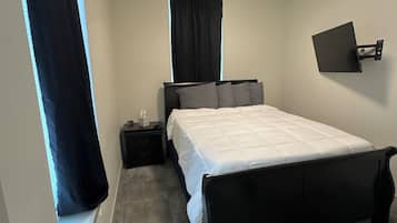 Una camera da letto con un letto, un comodino, una televisione a parete e una finestra con tende.