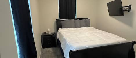 Un dormitorio con cama, mesita de noche, televisor en la pared y ventana con cortinas.