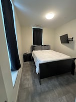 Un dormitorio con cama, mesita de noche, televisor en la pared y ventana con cortinas.