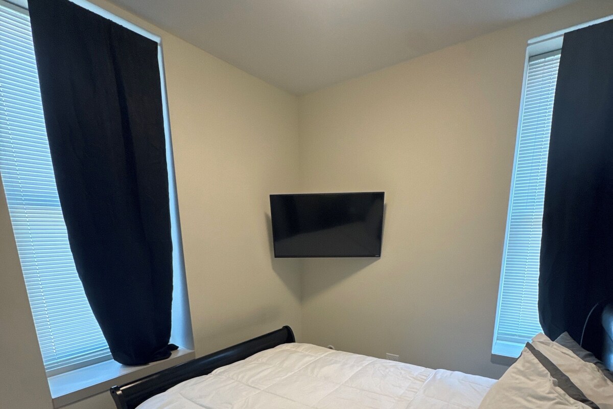 A bedroom with a bed, a flat-screen TV mounted on the wall, and windows with blinds.
