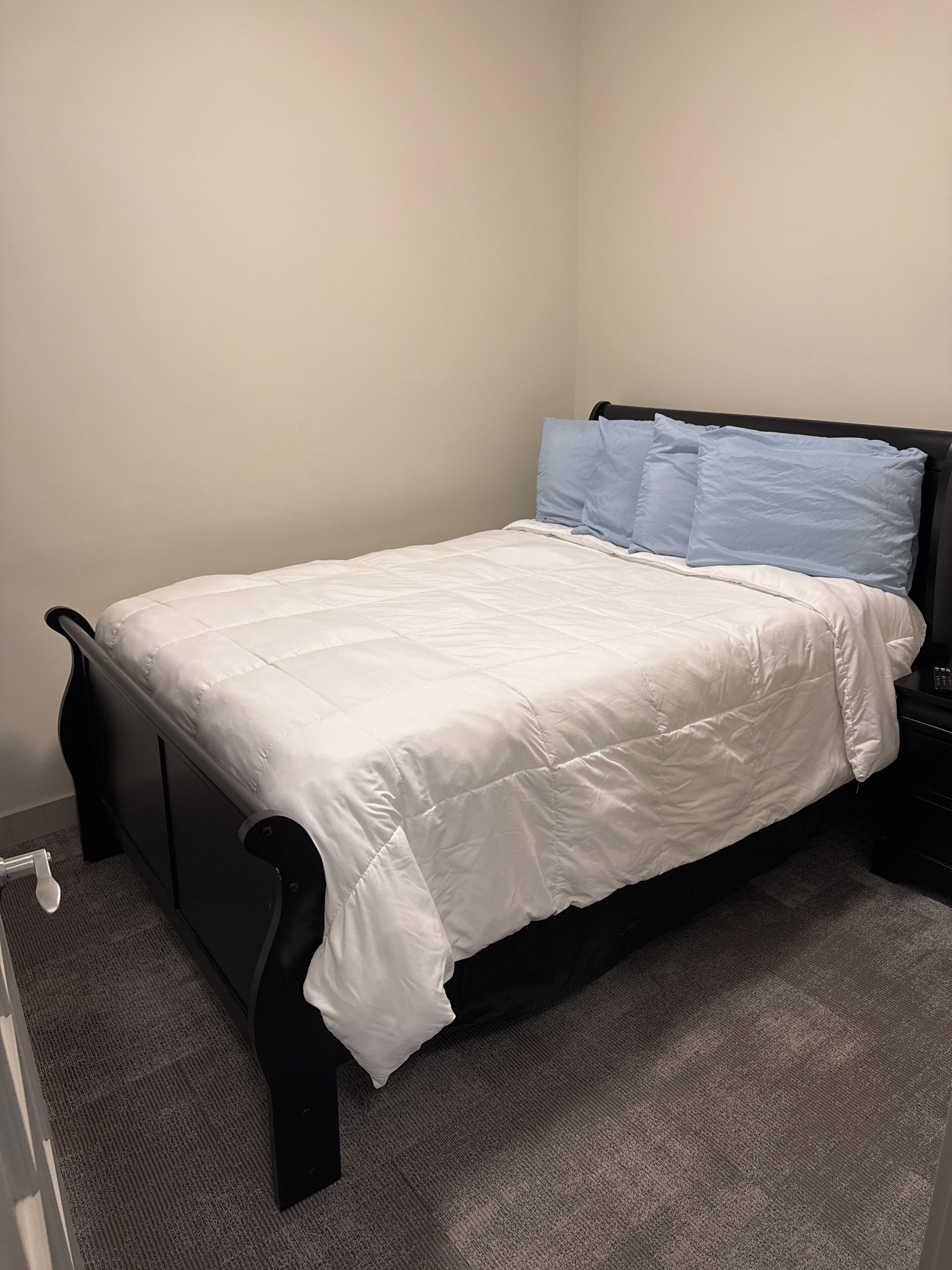 A single bed with a white duvet and two blue pillows, a black headboard, and a gray carpeted floor.