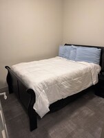 A single bed with a white duvet and two blue pillows, a black headboard, and a gray carpeted floor.