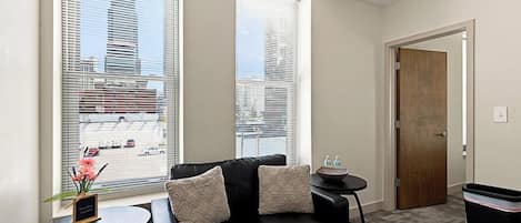A living room with a black leather sofa, two side tables, a small potted plant, and a view of a cityscape through large windows.